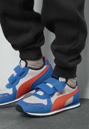 Baskets bleues avec des accents blancs et rouges, dotées de bandes Velcro, en matériaux en daim et synthétiques, et d'une semelle en caoutchouc noir. Pantalon noir uni visible.