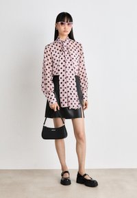 Langermet rosa bluse med svarte prikker, knyttet i kragen. Svart skinn mini-skjørt, svart platåsko, og en liten svart håndveske.
