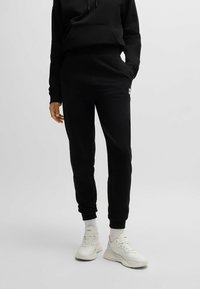 Zwarte sweatshirt met een voorzak en trekkoordcapuchon, gecombineerd met tapered zwarte joggers met een wit logo, en witte sneakers.