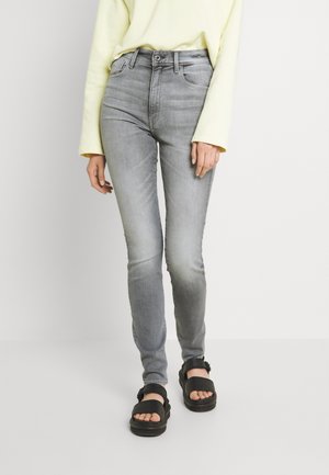 Osoba ubrana w jasnożółtą bluzkę z długim rękawem, wysokie, wyblakłe szare obcisłe dżinsy i czarne sandały z klamrami stojąca na białym tle.