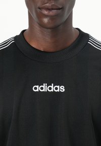 Fekete sportpóló bordázott nyakkivágással, három fehér csíkkal a vállakon és fehér "adidas" logóval a középen.