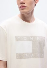 Μπλουζάκι με κοντά μανίκια σε κρεμ απόχρωση, με υφασμένο μπεζ γραφικό που γράφει "TOMMY" πάνω από το "JEANS", στρογγυλή λαιμόκοψη και απαλό βαμβακερό ύφασμα.