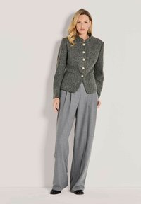 Vrouw in een grijze getextureerde knoopjasje met een staande kraag en brede lichtgrijze geplooide broek tegen een effen achtergrond.