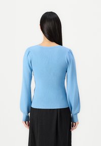 Suéter azul claro com textura acanalada, mangas longas e bufantes e corpo ajustado. Textura de malha suave, decote redondo, combinado com uma saia preta.