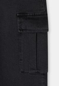 Gros plan sur un tissu en denim noir montrant une poche à rabat rectangulaire avec des coutures visibles et une couture verticale sur le côté droit.