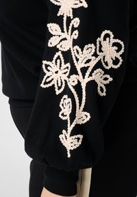 Zwarte trui met gestructureerde, crèmekleurige bloemrijke borduursels op de mouw. Voorzien van pofmouwen en een zachte stofafwerking.