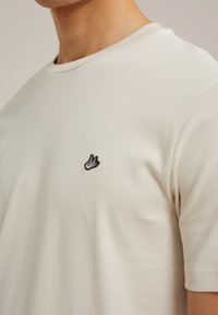 Světle béžové bavlněné tričko s kulatým výstřihem a krátkými rukávy. Na hrudi je malé černé vyšívané logo.