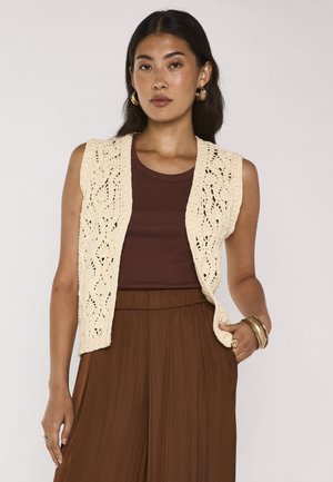 Woman with long dark hair wearing gold hoop earrings, cream crochet vest, brown sleeveless top, and brown pleated pants with hand in pocket.