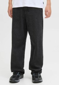 Personne portant un jean en denim noir ample et des chaussures de sport noires, se tenant devant un fond clair.