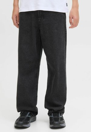 Personne portant un jean en denim noir ample et des chaussures de sport noires, se tenant devant un fond clair.