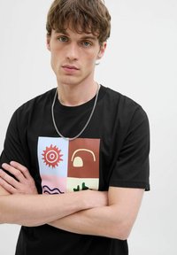 Schwarz Baumwoll-T-Shirt mit einem bunten Blockdesign mit Sonne, Welle und abstrakten Formen; runder Ausschnitt und kurze Ärmel. Silberne Kettenaccessoires.