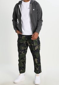 Sudadera gris con cremallera, camiseta blanca, pantalones cargo de camuflaje y zapatillas blancas. La sudadera tiene cordones y un pequeño logo. Los pantalones cargo tienen bolsillos.