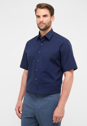 Man in een marineblauw korte mouwen overhemd met een borstzak en lichtblauwe broek voor een effen witte achtergrond.