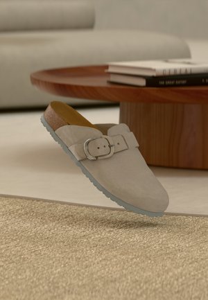 Beige slip-on træsko med en sølvspænde svæver over et tekstureret beige tæppe, et rundt træbord og stablede bøger i en neutral stue.