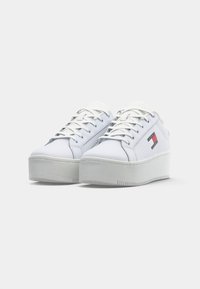 Biele platformové tenisky s hrubými podrážkami, bielymi šnúrkami a malým červeným, bielym a modrým logom Tommy Jeans na strane.