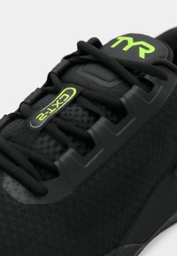 Zapatilla deportiva negra con parte superior texturizada, acentos de goma y marca en verde. Incluye un sistema de cordones tradicional para un ajuste seguro.
