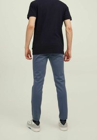 Hombre con camiseta negra, pantalones azules y zapatillas blancas de pie de espaldas frente a un fondo claro y sencillo.