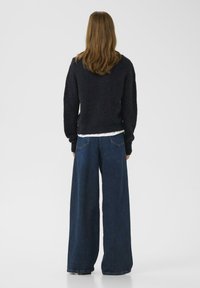 Přes oversized černý pletený svetr s kulatým výstřihem, v kombinaci s volnými džíny z tmavého denimu, které mají zadní kapsy a surový lem.