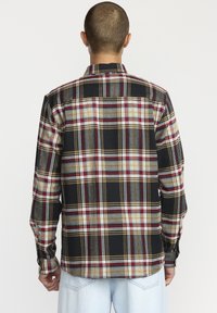 Langarmshirt met een zwart, rood, geel en grijs geruit patroon. Kenmerken zijn een knoopsluiting bij de kraag en gewatteerde mouwen, gemaakt van geweven stof.