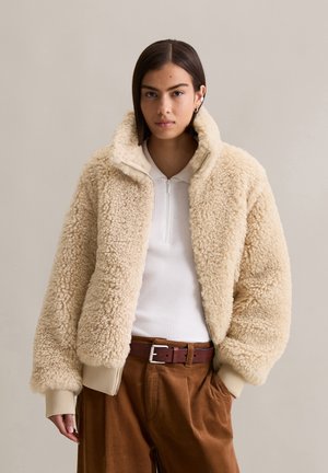 Vrouw in een beige sherpa jas over een witte ribgebreide rits-top en hoogstaande bruine broek met een leren riem, staand tegen een effen achtergrond.