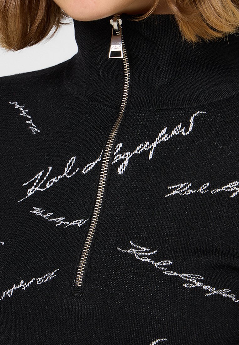 Pull noir à col montant, avec une fermeture éclair argentée et un design de script blanc sur l'ensemble. Texture tricotée, coupe près du corps.