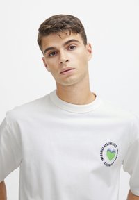 Hvid bomulds t-shirt med en afslappet pasform, der har et cirkulært grafisk motiv i blå og grøn på venstre brystområde. Glat tekstur.