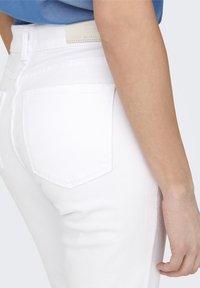 Person som bär vita denimjeans och en blå topp, visad från sidan med fokus på bakfickan och armen.