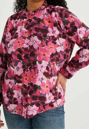 Vrouw die een blouse met knoopjes draagt met een bloemenpatroon in roze, paars en rood, en een blauwe spijkerbroek, met de hand op de heup.