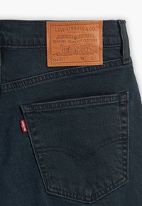Tmavé džínové kalhoty s hnědým koženým štítkem na pasu označeným "Levi Strauss & Co.". Červená etiketa na zadní kapse, podrobné švy.