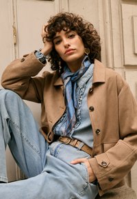 Jachetă maro din piele de căprioară, cămașă din denim bleu deschis cu volane, blugi cu talie înaltă și eșarfă albastră cu dungi portocalii. Cerceii aurii completează ținuta.