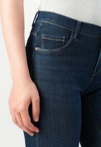 Close-up of a person’s hand resting beside dark blue denim jeans with visible stitching and front pocket detail.