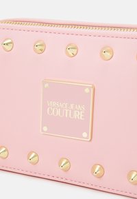 Różowy portfel z materiału syntetycznego z ozdobnymi złotymi ćwiekami i prostokątną plakietką z logo. Zamknięcie na suwak na górze z ozdobnym przeszyciem.