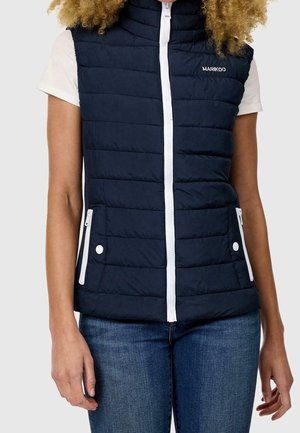 Femme portant un gilet matelassé bleu marine avec une fermeture éclair blanche et des poches, une chemise blanche à manches courtes et un jean bleu.