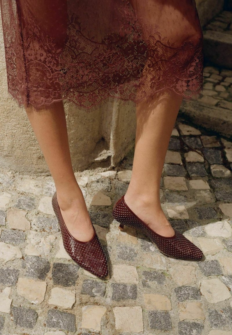 Nohy v bordó krajkové sukni se stejnobarevnými texturovanými botami na nízkém podpatku se špičatou špičkou stojí na dlážděné dlažbě.