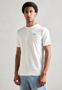 Jauns vīrietis ar īsiem, lokainiem matiem, tērpies baltā Marc O'Polo Denim t-kreklā un gaiši zilās džinsās, stāv pretī vienkāršam gaišam foniem.