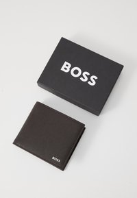 Portafoglio in pelle marrone con finitura testurizzata, dotato di un logo bianco "BOSS". È posizionato accanto a una scatola nera opaca etichettata "BOSS."