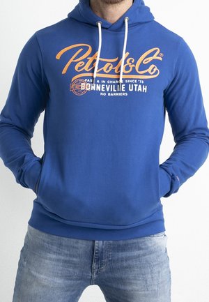 Uomo che indossa una felpa con cappuccio blu con logo arancione "Petrol & Co" e cordini bianchi, abbinata a jeans azzurri chiari, con le mani in tasca.