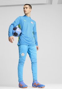Puma sportoutfit in lichtblauw, met een rits bovenkant en bijpassende broek. Opmerkelijke groene accenten, gestructureerde stof en clublogo. Blauwe voetbal inbegrepen.