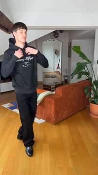 Νεαρός που φοράει μαύρο φούτερ Tommy Hilfiger, που ρυθμίζει το φούτερ του σε ένα μοντέρνο σαλόνι με πορτοκαλί καναπέ και μεγάλο γλαστρικό φυτό.