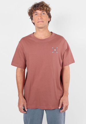 Homme aux cheveux bouclés porte un t-shirt marron à manches courtes avec un petit motif floral sur la poitrine gauche et un pantalon gris, debout sur un fond blanc.