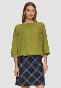 Olijfgroene, gestructureerde blouse met driekwartmouwen, gecombineerd met een zwart-blauwe ruitrok. De outfit heeft een losse, ontspannen pasvorm.