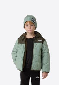 Padjaga isle sinist rohelist jope tumedate roheliste detailidega, millel on kapuuts, esist lukku ja brändimärgistusega plaastrid. Sobitatud samas toonis kootud mütsiga.