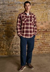 Young man standing against a brick wall wearing a red plaid shirt, navy blue pants, and brown leather shoes with hands in pockets.