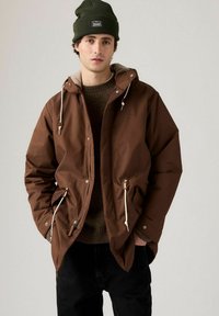 Brun vattentät jacka med huva, dragsnoddar och två framfickor. Buren över en brun tröja, i kombination med mörka jeans.