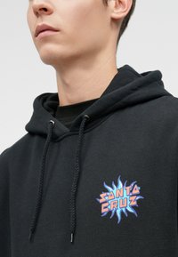 Fiatal személy fekete kapucnis pulóverben, a mellkasán egy színes "Santa Cruz" lánglogóval és lelógó zsinórokkal.