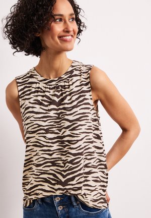 Femme aux cheveux bouclés portant un haut sans manches à imprimé zèbre et un jean bleu, souriante et regardant sur le côté sur un fond uni.