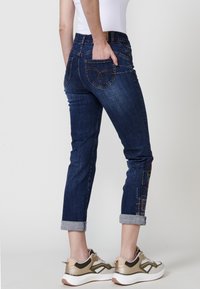 Temno modri denim kavbojki s skrajšanim krojem, zavitimi manšetami in barvitim vezenjem po nogi. Imajo zadnje žepe s ukrivljenim šivom.