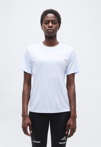 Γυναίκα που στέκεται μπροστά φορώντας αθλητικό μπλουζάκι κοντομάνικο ανοιχτού μπλε χρώματος και μαύρο κολάν  με ανακλαστικά λογότυπα Adidas.