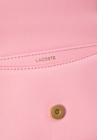 Rožinės odos gaminys su auksiniu folijos „LACOSTE“ logotipu ir apvaliu auksiniu snapeliu. Lygi tekstūra su siūlėmis.