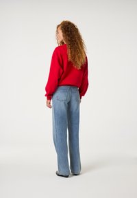 Roter übergro�ßer Sweatshirt mit überschnittenen Schultern, kombiniert mit hellblauen weiten Jeans. Das Model hat lockige Haare und trägt dezente Hängeringe.